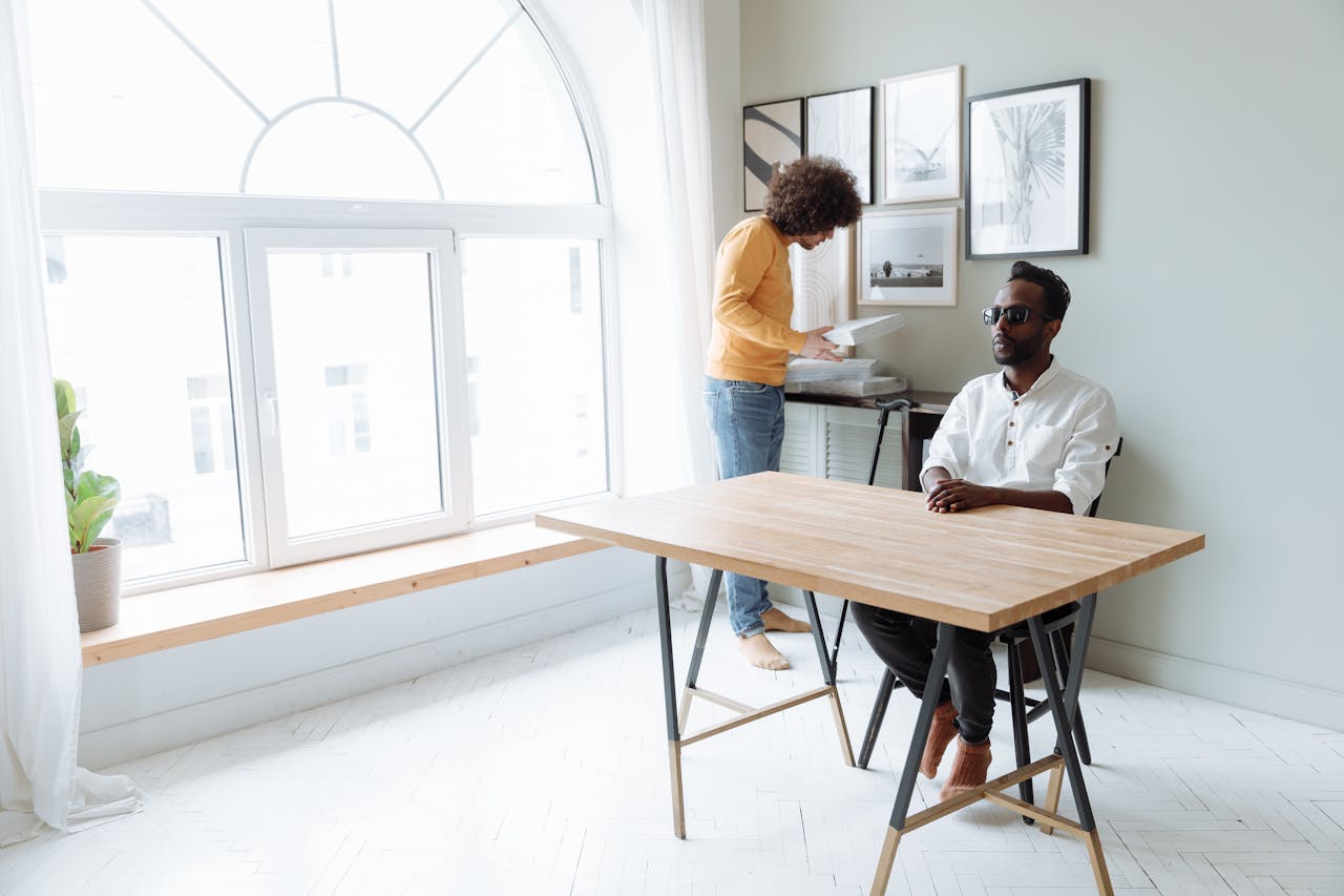 A visually impaired man sitting at a table while a mentor prepares materials in a bright room.
